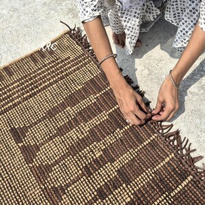 May include: A handwoven rectangular rug with a geometric pattern in shades of brown and beige. The rug has a fringed edge and is being handled by a person wearing a patterned top and bracelets.