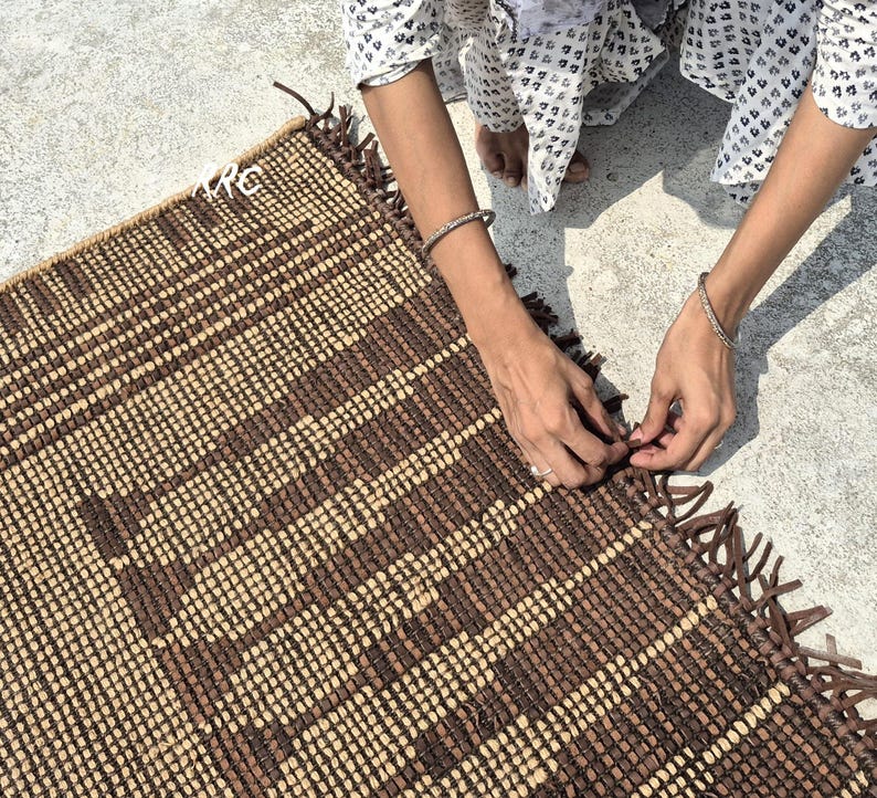 May include: A handwoven rectangular rug with a geometric pattern in shades of brown and beige. The rug has a fringed edge and is being handled by a person wearing a patterned top and bracelets.