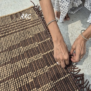 May include: A rectangular woven rug with a brown and beige pattern. The rug has a fringed edge and the letters "RRC" are visible in the upper left corner. The rug is being handled by a person.
