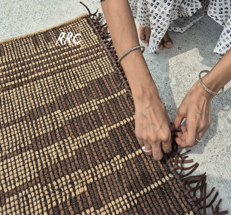 May include: A rectangular woven rug with a brown and beige pattern. The rug has a fringed edge and the letters "RRC" are visible in the upper left corner. The rug is being handled by a person.