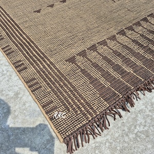 May include: A woven brown and tan rug with a geometric pattern. The rug features a repeating pattern of vertical stripes and triangles. The edge of the rug has a fringe detail. The rug is made of a natural fiber.
