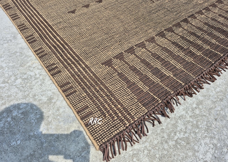 May include: A woven brown and tan rug with a geometric pattern. The rug features a repeating pattern of vertical stripes and triangles. The edge of the rug has a fringe detail. The rug is made of a natural fiber.
