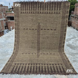 May include: A large, rectangular woven rug with a geometric pattern in shades of brown and tan. The rug features a repeating design of triangles and vertical lines, with a fringed edge. The letters "RRC" are visible in the upper left and lower right corners.