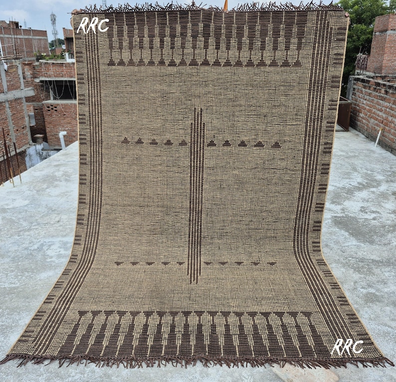 May include: A large, rectangular woven rug with a geometric pattern in shades of brown and tan. The rug features a repeating design of triangles and vertical lines, with a fringed edge. The letters "RRC" are visible in the upper left and lower right corners.