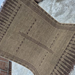 May include: A rectangular, woven rug in shades of brown and tan. The rug features a central design of horizontal lines and triangles, with a fringed edge. The letters "RRC" are visible in the corners.