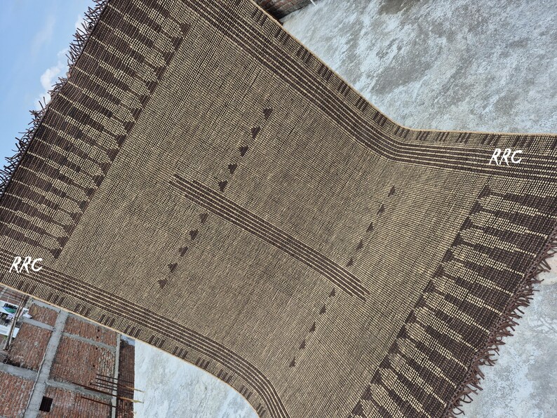 May include: A rectangular, woven rug in shades of brown and tan. The rug features a central design of horizontal lines and triangles, with a fringed edge. The letters "RRC" are visible in the corners.