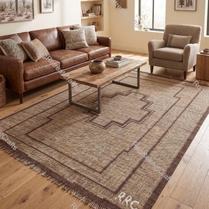 Peut inclure: Un tapis rectangulaire à la texture tissée dans des tons de beige et de brun. Le tapis présente un motif de bordure géométrique marron foncé et inclut les lettres RRC brodées dans le coin. Il est fini avec des franges courtes aux extrémités.