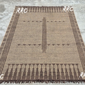 May include: Rectangular brown woven rug with a geometric design. The rug features a central vertical stripe and decorative borders with fringe. The corners of the rug have the letters "RRC" in white.