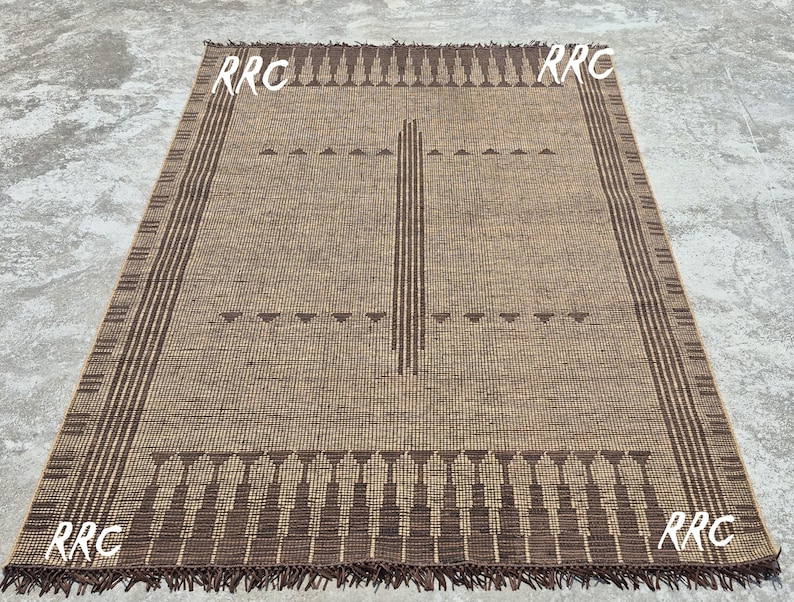 May include: Rectangular brown woven rug with a geometric design. The rug features a central vertical stripe and decorative borders with fringe. The corners of the rug have the letters "RRC" in white.