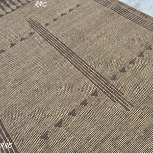 May include: A woven rug with a tan background and dark brown geometric patterns. The design includes vertical stripes and triangle shapes. The rug has a textured appearance and a rectangular shape, suitable for home decor.