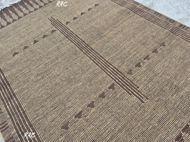 May include: A woven rug with a tan background and dark brown geometric patterns. The design includes vertical stripes and triangle shapes. The rug has a textured appearance and a rectangular shape, suitable for home decor.
