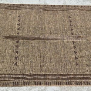 May include: A rectangular brown rug with a geometric design. The rug features a central pattern of horizontal lines and triangles, with decorative fringe on the edges. The letters "RRC" are visible in the corners.