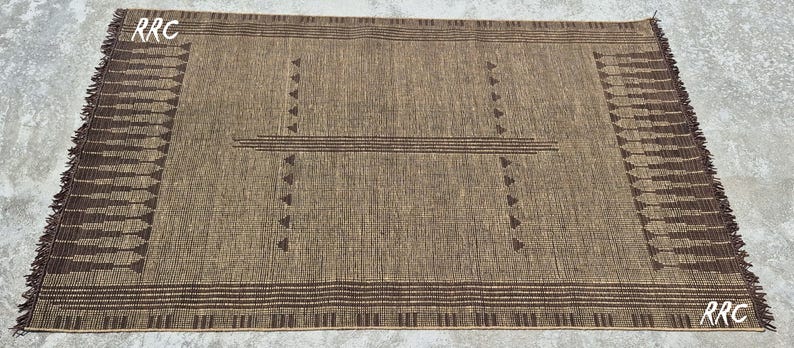 May include: A rectangular brown rug with a geometric design. The rug features a central pattern of horizontal lines and triangles, with decorative fringe on the edges. The letters "RRC" are visible in the corners.
