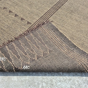 May include: A woven rug with a geometric pattern in shades of brown and beige. The rug features diagonal stripes and small triangular shapes. The underside of the rug is visible, showing a darker brown color with stitched details and the letters "RRC".
