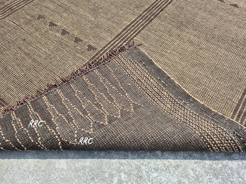 May include: A woven rug with a geometric pattern in shades of brown and beige. The rug features diagonal stripes and small triangular shapes. The underside of the rug is visible, showing a darker brown color with stitched details and the letters "RRC".