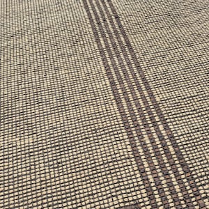 May include: Close-up of a woven rug with a textured surface. The rug has a beige background with a dark brown grid pattern. Three vertical dark brown stripes run across the rug.