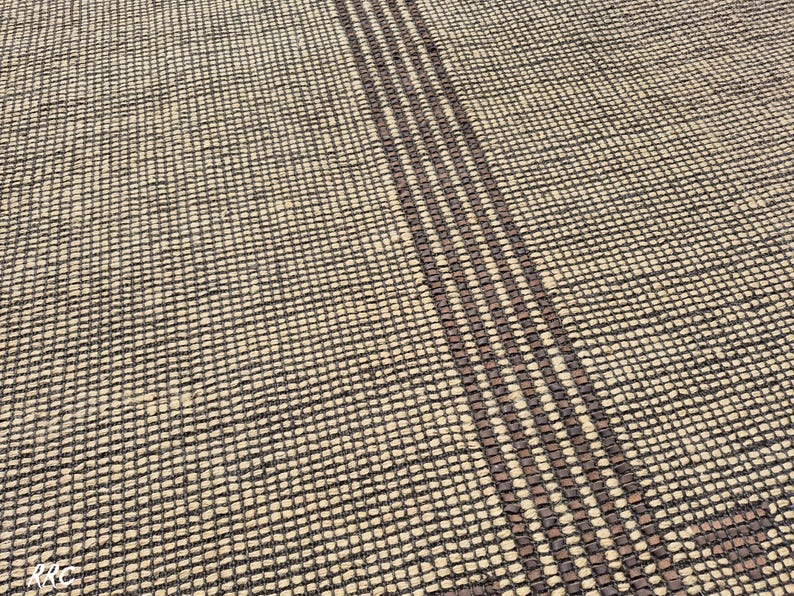 May include: Close-up of a woven rug with a textured surface. The rug has a beige background with a dark brown grid pattern. Three vertical dark brown stripes run across the rug.