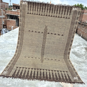 May include: A rectangular brown and tan woven rug with a geometric pattern. The rug features dark brown vertical stripes and decorative elements along the edges. The corners of the rug have the text "RRC".