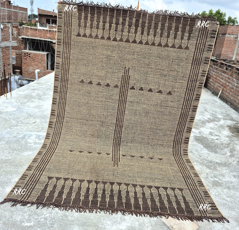 May include: A rectangular brown and tan woven rug with a geometric pattern. The rug features dark brown vertical stripes and decorative elements along the edges. The corners of the rug have the text "RRC".