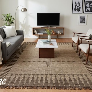 May include: A neutral-toned area rug with a geometric pattern and fringe. The rug is in a living room setting with a gray sofa, white chairs, and a wooden coffee table. The rug's dimensions are approximately 8 x 10 feet.
