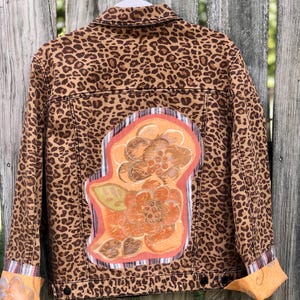 May include: A brown leopard print jacket with a floral design on the back. The jacket has a classic collar, button closures, and orange cuffs. The floral design features orange and brown flowers with a pink border.