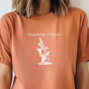 May include: Rust-colored t-shirt with the phrase "Knowledge is Power" above a white illustration of a microscope. The shirt has short sleeves and a crew neck.