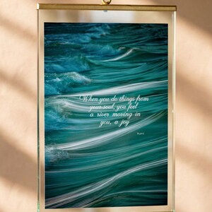 May include: A framed print with a teal ocean wave image and the quote "When you do things from your soul, you feel a river moving in you, a joy" by Rumi. The frame is gold-coloured and suspended by a gold chain.
