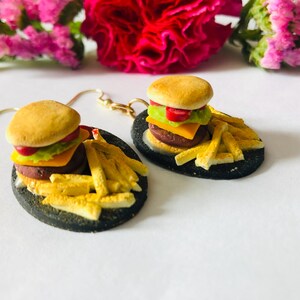 May include: A pair of novelty earrings featuring miniature burger and fries designs. Each earring has a small burger with a bun, patty, cheese, lettuce, and tomato, served with a side of golden fries on a black oval plate.