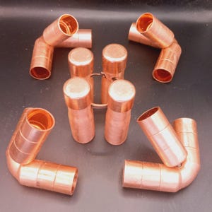 Può includere: Raccordi idraulici in rame, tra cui gomiti e tubi tappati, sono disposti su uno sfondo scuro. I raccordi hanno una finitura metallica lucida e sono progettati per collegare tubi in sistemi idraulici.