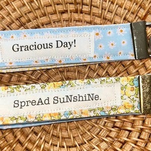 May include: Two fabric keychains with metal hardware. One is light blue with white daisies and the text "Gracious Day!". The other is floral with the text "SpreAd SuNshiNe."
