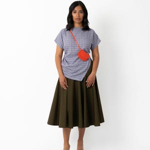 May include: A woman wearing a blue and white plaid top, an olive green skirt, and black heels. A small, bright orange purse is worn across the body. The outfit is set against a white background.