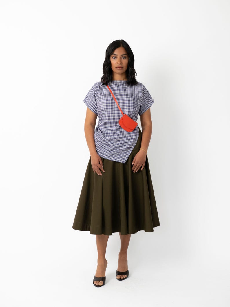 May include: A woman wearing a blue and white plaid top, an olive green skirt, and black heels. A small, bright orange purse is worn across the body. The outfit is set against a white background.