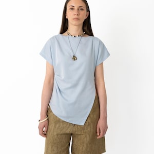 May include: A light blue top with short sleeves and an asymmetrical hemline, paired with olive green shorts. The outfit is accessorized with a necklace and a bracelet. The model is standing in front of a white background.