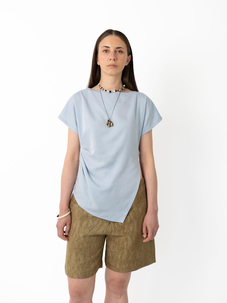 May include: A light blue top with short sleeves and an asymmetrical hemline, paired with olive green shorts. The outfit is accessorized with a necklace and a bracelet. The model is standing in front of a white background.