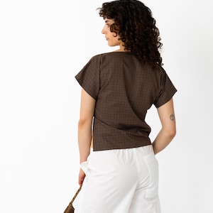 May include: A brown plaid short-sleeve top and white pants. The top has a V-neck and a subtle checkered pattern. The pants are wide-legged and high-waisted. A small woven bag is held in the hand.