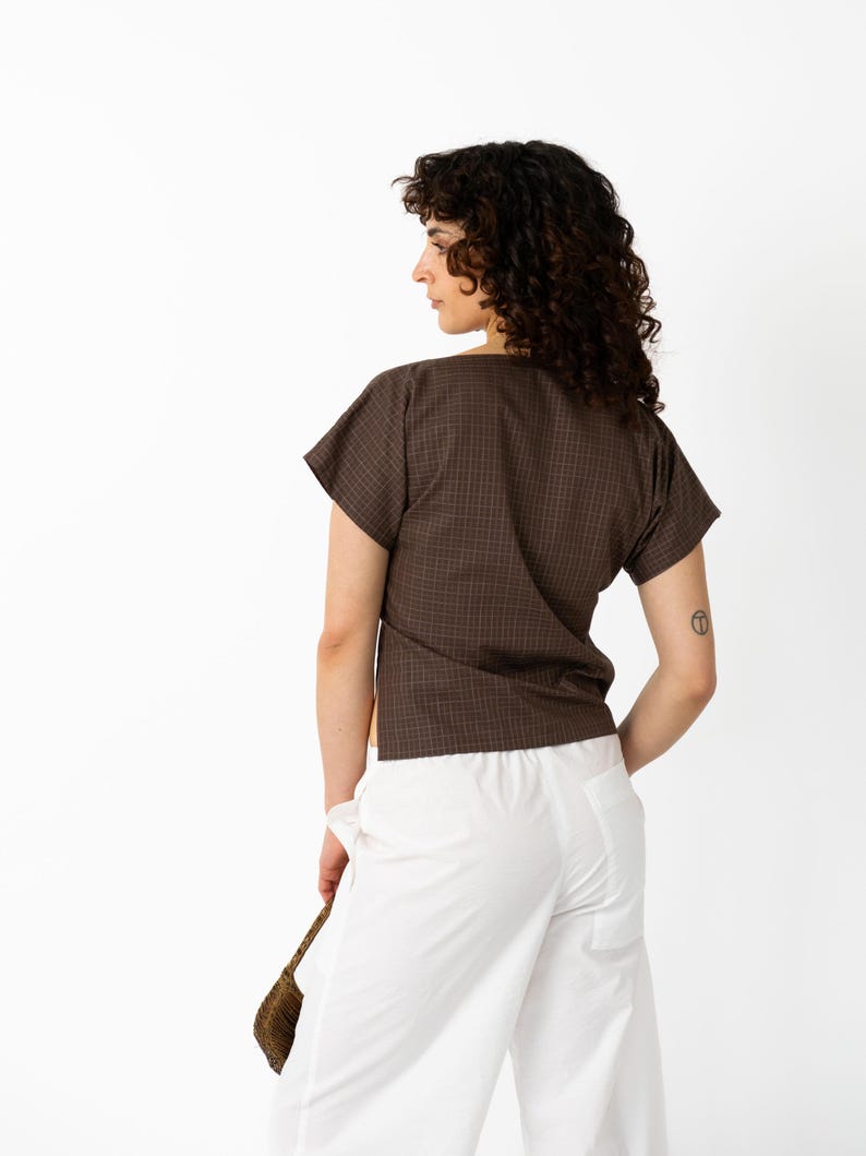 May include: A brown plaid short-sleeve top and white pants. The top has a V-neck and a subtle checkered pattern. The pants are wide-legged and high-waisted. A small woven bag is held in the hand.