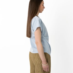 May include: A light blue short-sleeved top with a draped front detail. Paired with brown shorts featuring a textured pattern. A white bracelet is visible on the wrist. The outfit is set against a plain white background, showcasing the clothing.