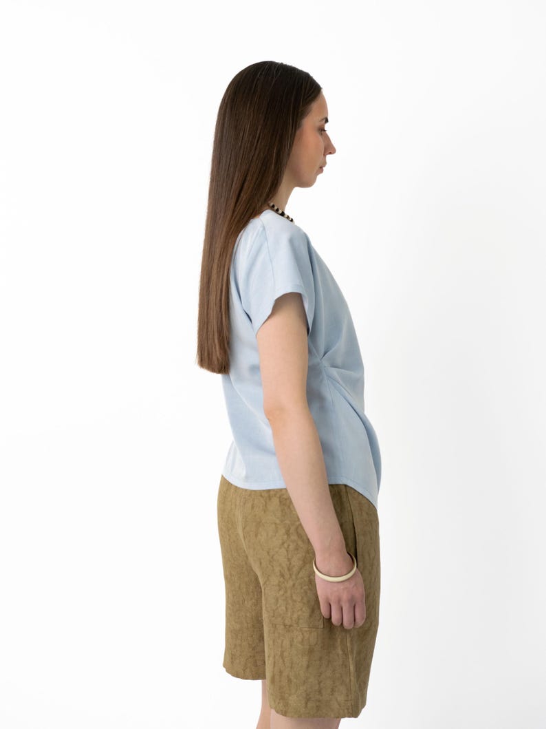 May include: A light blue short-sleeved top with a draped front detail. Paired with brown shorts featuring a textured pattern. A white bracelet is visible on the wrist. The outfit is set against a plain white background, showcasing the clothing.