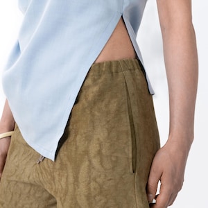 May include: A light blue sleeveless top with a side slit is paired with olive green pants. The pants have a textured pattern and a drawstring waist. The outfit is simple and casual.