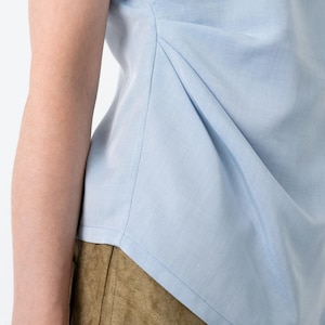May include: A light blue top with a draped design. The top features a gathered detail on the side, creating a textured effect. The fabric appears to be a lightweight material, suitable for warm weather. The top is paired with olive-green pants.