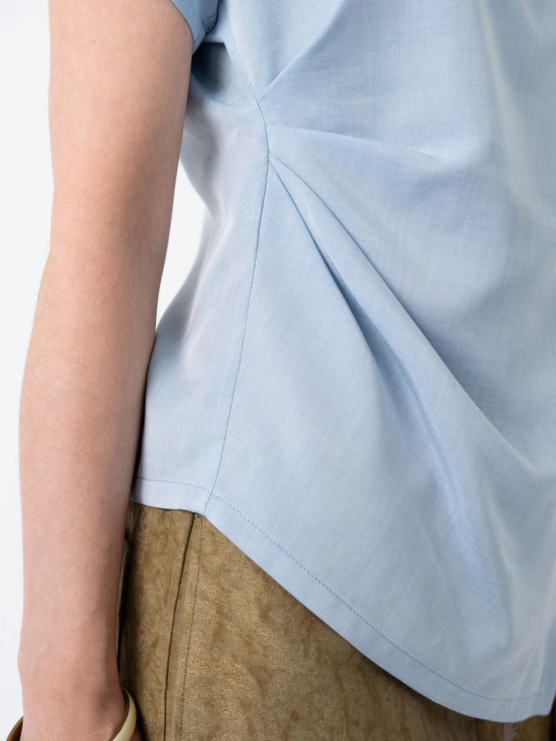 May include: A light blue top with a draped design. The top features a gathered detail on the side, creating a textured effect. The fabric appears to be a lightweight material, suitable for warm weather. The top is paired with olive-green pants.