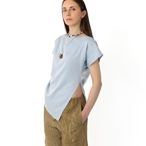 May include: A light blue top with a unique asymmetrical design, paired with olive green shorts. The top has short sleeves and a split side. A beaded necklace with a pendant is also visible.