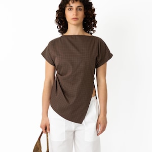 May include: A brown, short-sleeved top with a square neckline and a unique asymmetrical design. The top is paired with white pants. A small, woven brown handbag is held in the hand.
