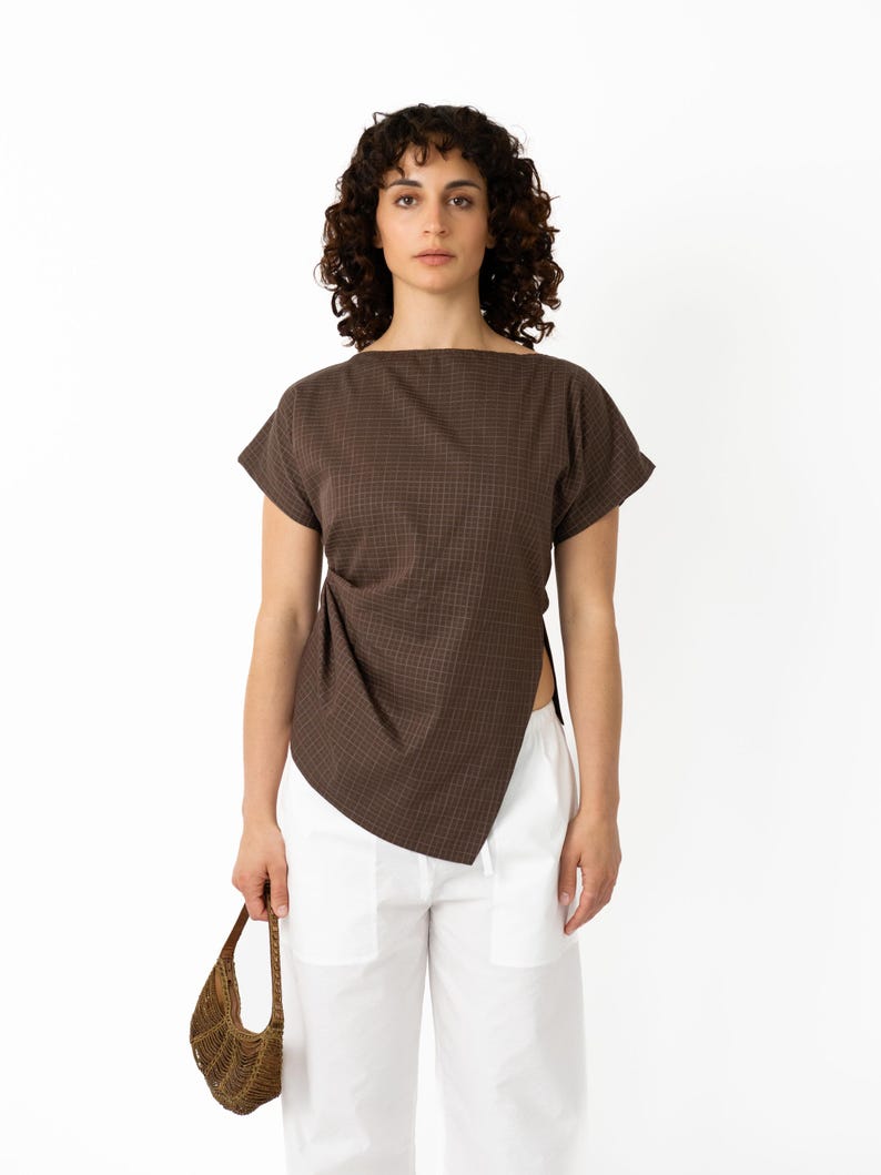 May include: A brown, short-sleeved top with a square neckline and a unique asymmetrical design. The top is paired with white pants. A small, woven brown handbag is held in the hand.