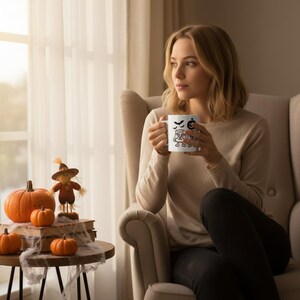 Puede incluir: Una taza de cerámica blanca con un diseño de Halloween que presenta murciélagos, una calabaza y la palabra "Spooky". La taza es sostenida por una persona. Una pequeña mesa exhibe calabazas y un espantapájaros.