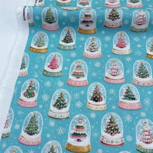 May include: A teal fabric featuring a repeating pattern of Christmas trees inside glass snow globes. The globes have gold-toned bases and are surrounded by white snowflake illustrations. The fabric has a white selvage edge with printed text.