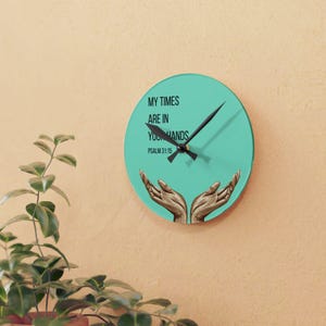 May include: A teal-faced wall clock with the text "MY TIMES ARE IN YOUR HANDS" and "PSALM 31:15". The clock has black hands and an illustration of two hands. The clock is mounted on a light beige wall. The clock is a decorative item.