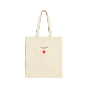 Könnte beinhalten: Cremefarbene Tragetasche mit langen Henkeln. Die Tasche zeigt den Text "GOD IS LOVE" über einem kleinen roten Herzen, mit dem Bibelvers Johannes 4:16 darunter. Die Tasche ist aus einem robusten Stoff gefertigt.