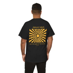 Könnte beinhalten: Schwarzes T-Shirt mit einem gelb-schwarzen Sonnenstrahlen-Design auf dem Rücken. Der Text "Jesus says" steht über dem Sonnenstrahlen-Design, darunter "I am the light of the world. John 8:12" in einfacher Schrift.