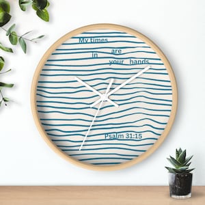 May include: A round wall clock with a light wood frame. The clock face features a cream-colored background with blue horizontal wavy lines. The text "My times are in your hands" and "Psalm 31:15" are printed on the face.